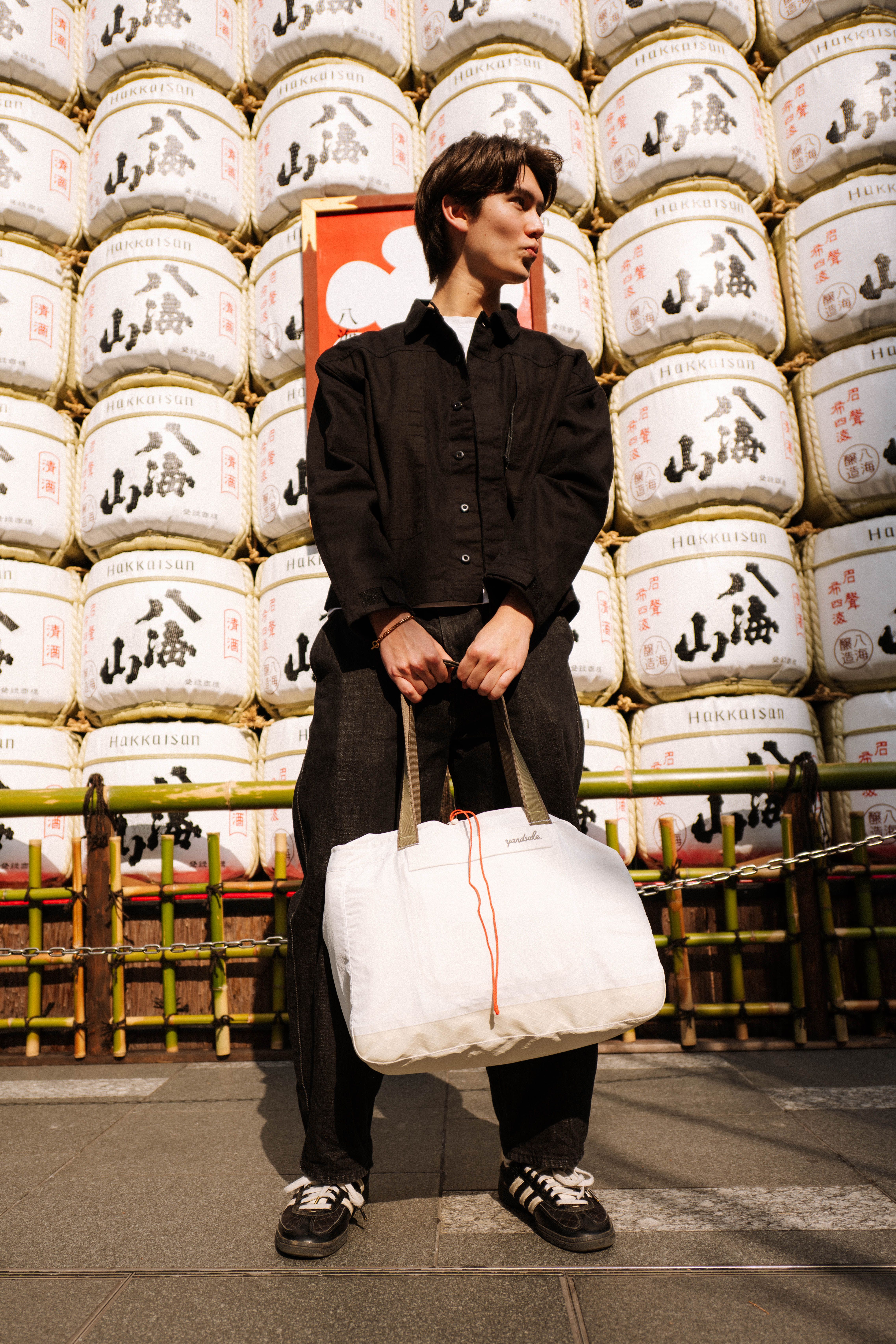 A man holding a 20L Day Bag in Tokyo in front of sake barrels
